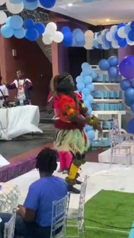 Here's a concise description of the video:

The video captures a festive event, likely a birthday party, indoors. Blue and white balloons are strung across the ceiling as decorations. A DJ set is present, with people dancing and enjoying the music. A person in traditional attire is dancing prominently. There's also a decorated area with what looks like a cake and gifts. People of various ages are present.