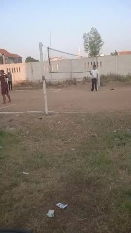 A group of people are playing volleyball on a dirt court. A white net is stretched across the court. The sky is clear and the sun is setting. There are buildings in the background.