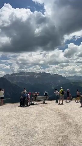The video shows a group of people gathered on a gravelly overlook, with a panoramic view of mountains under a cloudy sky. Several individuals are standing, some holding cameras or phones and appearing to take photos or videos. Others are seated on a bench. The people are dressed in casual, outdoor clothing, including hats and backpacks. The environment is a mountainous landscape with rocky peaks and lush green valleys visible in the distance. The sky is partly cloudy with dramatic white cumulus clouds against a blue background. The overall atmosphere appears to be one of tourism and appreciation of the natural scenery.