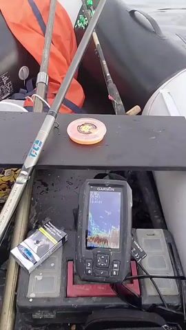 The video shows a first-person perspective from an inflatable boat on a large body of water, likely a lake or reservoir, with distant land and hills under a cloudy sky. The water is calm with gentle ripples.

Inside the boat, various fishing equipment is visible. An orange life vest is folded and placed on the side of the boat. Several fishing rods are set up, one leaning against the boat's side with "TCS" written on its handle. A yellow fishing float is also seen near the rods. A fishing net with a long handle is positioned, possibly ready for use. On a flat surface in the boat, an orange spool of fishing line is placed next to a Garmin fishfinder with its screen on. Oars are also visible along the boat's side.

The scene suggests an ongoing or upcoming fishing trip. The time of day appears to be daytime, with an overcast sky. The overall atmosphere is tranquil.