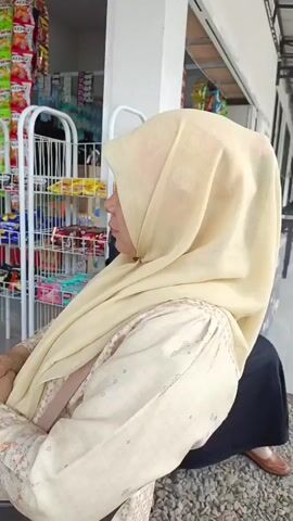 A woman wearing a light yellow hijab and a patterned light-colored blouse is seated facing left, with her back to the camera. She is sitting in front of a display rack filled with various packaged goods, predominantly snacks and drinks. The rack is white metal and has multiple shelves. Behind the woman and the rack is an open-fronted building with a white exterior and a grey overhead structure. The ground appears to be gravel. The woman's hands are clasped in her lap. The scene is brightly lit, suggesting it is daytime.