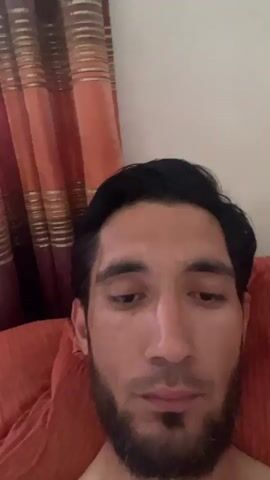 This is a selfie video of a man with dark hair and a beard. The man's face fills the majority of the frame. He is lying down, with an orange textured surface behind his head, likely a pillow or couch. Behind him and to the left, there are curtains that are orange and brown with gold stripes. The lighting suggests it is daytime. The man makes a series of facial expressions: he looks down, then his eyes widen in surprise, followed by a grimace, and finally a wink. His expression seems playful and lighthearted. There is no visible text in the video. The setting appears to be indoors, in a room with curtains, possibly a bedroom or living room.