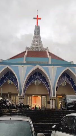 The video shows the exterior of a church with a prominent red cross at the top, illuminated against a cloudy sky. The church has a modern architectural design featuring large arched entrances with blue and white accents. Several cars are parked in front of the church, and a few motorcycles are visible on the left side of the frame. Steps lead up to the entrance, and a few figures can be seen inside the brightly lit doorway. The overall atmosphere appears calm and overcast.
