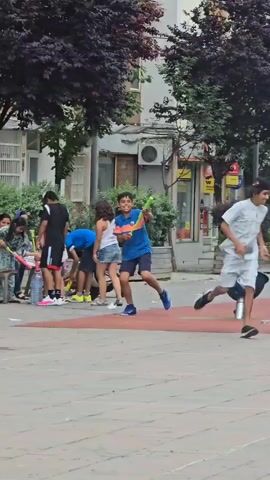A group of children are playing outside in a paved area. The child in the foreground, wearing a blue shirt and shorts, is holding a toy bat and appears to be swinging it. Other children are gathered around, some are holding similar toys. In the background, there are buildings, trees, and a park bench. The overall atmosphere suggests a casual outdoor activity.