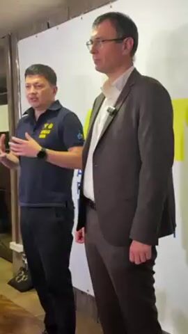 Two men are standing next to each other in front of a backdrop with yellow and blue coloring. The man on the left has short black hair, dark pants, and a dark blue polo shirt with yellow writing. He is wearing a watch on his left wrist and gesturing with both hands. The man on the right has short brown hair, glasses, a white button-down shirt, brown pants, and a brown blazer. He has a lavalier microphone clipped to his shirt. A third person is slightly visible on the left. The background is partially out of focus. The light in the room is soft.