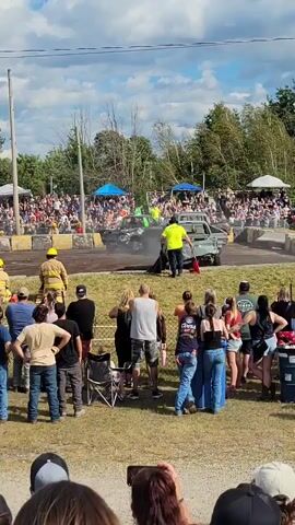 A demolition derby event is underway with several cars in the arena. A green car with heavy damage is spinning its wheels, emitting smoke. Two individuals in bright yellow safety vests are standing near the green car, one holding what appears to be a fire extinguisher. Two individuals in firefighter gear are also present, observing the situation. A crowd of spectators is gathered behind a barrier, watching the event. The environment is outdoors with a cloudy sky and trees in the background.