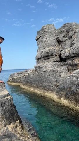 A young man in orange swim trunks jumps off a rocky cliff into clear, turquoise water. The cliff is made of gray rock, and a second, larger cliff rises from the other side of the water. The sky is blue with a few white clouds. The man jumps forward, spreads his arms, and enters the water feet-first, creating a splash. He then surfaces and swims.