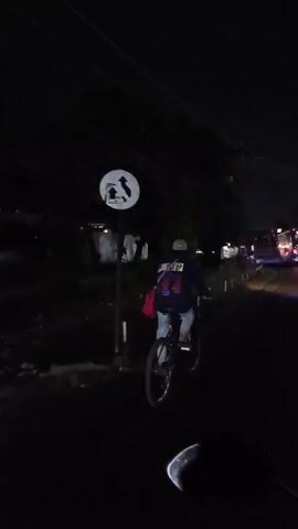 The video shows a person riding a bicycle at night on a paved road. The rider is wearing a dark blue shirt with a red and white logo on the back, jeans, and a cap. A red bag is slung over their left shoulder. The bicycle is black with red accents on the rims. The road is dark with some faint streetlights visible in the distance, suggesting an urban or suburban environment. Trees and buildings are dimly visible along the side of the road. The perspective is from behind and slightly to the right of the cyclist, indicating the video is likely being filmed from another moving vehicle. The overall atmosphere is dark and nocturnal.