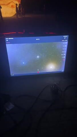 The video shows a dimly lit outdoor area at night. In the foreground, a large screen displays an image of a starfield. A computer mouse and cables are visible on the ground in front of the screen. In the background, several large, inflatable spheres resembling planets are illuminated. Silhouettes of people can be seen near these spheres. Red laser light or LED lighting is also present in the background, creating lines of light on the ground. The overall scene appears to be an outdoor exhibition or event related to space or astronomy.