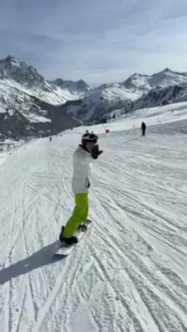 A person snowboards down a snowy mountain, wearing a white coat and bright green pants, with goggles and a helmet. Their arms are initially raised. In the background, there are snow-capped mountains under a partly cloudy sky. Other people are also snowboarding or skiing on the slope. The snow is covered with tracks.