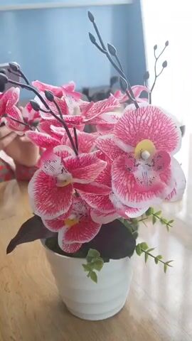A potted artificial orchid with pink and white flowers is shown. The orchid is in a white pot and is placed on a wooden table. In the background, a person's arm can be seen, and a glass with red liquid is visible. The background is blurred, suggesting a shallow depth of field. The lighting is bright, highlighting the flowers.