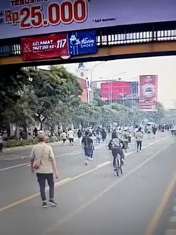 The video shows a street scene with multiple people walking and running. A large billboard with red text that appears to say "Rp 25.000" is visible at the top. There are trees and buildings along the street. Some people are dressed in casual clothing, while others are wearing more athletic attire. One person is riding a bicycle. There are also several bicycles parked on the side of the street. The overall impression is of a public space with people engaging in outdoor activities.