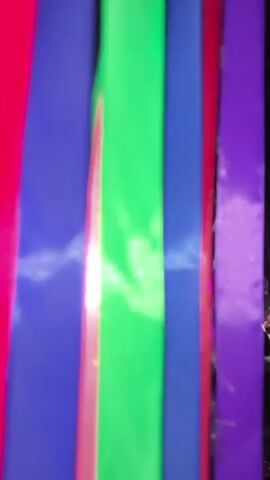 The video opens with out-of-focus shots of green and white light. It then transitions to a nighttime outdoor scene with many people milling about and seated. Long, colorful fabric streamers are hanging from overhead wires, illuminated by various colored lights. The ground is covered with grass or dirt. There appears to be some sort of gathering or event taking place. People are dressed casually. The overall atmosphere is festive and relaxed.
