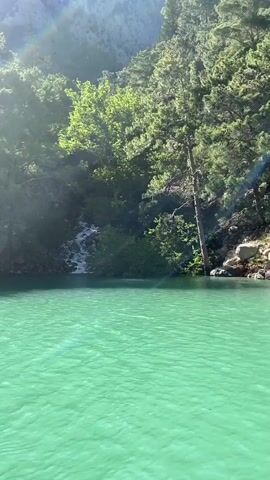 A still, wide shot captures a picturesque natural scene dominated by a vibrant turquoise body of water, likely a lake or river, filling the foreground. On the left side, a small waterfall cascades down a rocky, tree-covered slope, emptying into the main body of water. The trees are lush and green, indicating a healthy, natural environment. Sunlight filters through the foliage, creating bright patches and a lens flare effect on the left side of the frame. The background consists of steep, forested mountainsides, adding to the grandeur of the landscape. The water's distinct color suggests a mineral-rich composition, common in regions like Akseki, Turkey. There are no people, animals, or man-made structures visible, emphasizing the untouched beauty of the natural setting. The time of day appears to be sunny, showcasing the clear water and verdant surroundings.