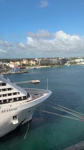 The video shows the front side of the MSC Seaside cruise ship docked in port on a partly cloudy day. The ship is white with multiple decks visible, and its name is written clearly on the side.  Across the water, a cityscape with buildings, including hotels, can be seen. The water in the harbor is calm and turquoise blue. A small dock and some boats are also visible near the shore.