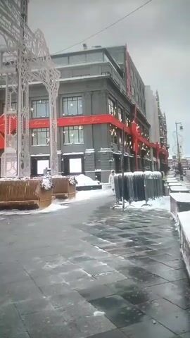 The video shows a snowy cityscape, featuring a large, grey building decorated with a red ribbon and bow, along with the words "Happy New Year". In front of the building is a pedestrian area covered in snow and ice, with decorative seating arrangements made of light-colored wood and what appears to be a white metal gazebo-like structure, also coated in snow. A person wearing a light-colored coat is walking in the distance. The sky is overcast and the overall impression is one of a quiet, wintry scene.