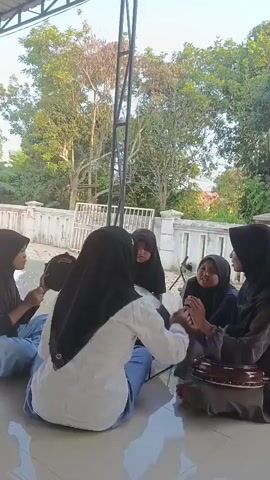 A group of five young females wearing hijabs are sitting in a circle on the floor outdoors. Some of them are holding small percussion instruments. The setting appears to be a covered porch or patio with trees and a white fence in the background. The lighting suggests it is daytime.