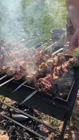 The video shows skewers loaded with meat grilling over hot coals in a portable black grill. The person handling the skewers is wearing a floral patterned shirt and orange gloves. Plumes of white smoke rise from the grill. A silver bucket sits on the grass in the background. The grill is positioned on a patch of sandy soil and grass, indicating an outdoor setting. The meat on the skewers varies in color, some pieces browned and cooked, others appearing lighter and less done.