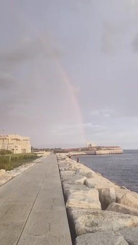 A faint rainbow arcs across a cloudy sky above a waterfront promenade. The promenade, made of concrete pavers, stretches into the distance, bordered on the right by a wall of large, light-colored rocks. On the left, a low, white building with a flat roof sits behind a strip of green vegetation. The sea is visible to the right of the rocky embankment. Two figures can be seen walking on the promenade in the distance.