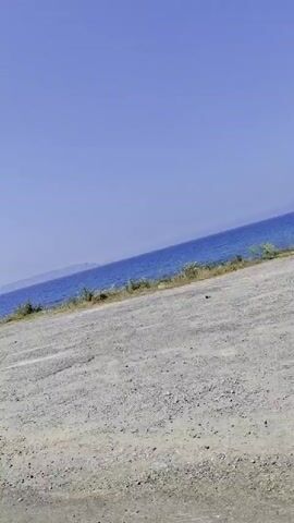 The video shows a clear, blue sea under a clear, blue sky. The video appears to have been taken from a moving vehicle traveling along a road with a gray, pebbled shoulder. A roadside barrier runs along the shoulder of the road.  Vegetation can be seen between the barrier and the sea. In the distance, a landmass is faintly visible on the horizon.