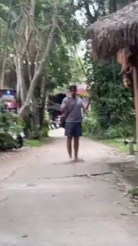 The video shows a man walking on a paved path. The man has a mustache and is wearing a gray t-shirt, patterned shorts, and sandals. He is carrying a bag over his shoulder and holding a cell phone in his hand. The path is surrounded by green trees and foliage. There is a thatched-roof structure to the right and a building and parking area in the background. The man pauses and turns slightly to face the camera.