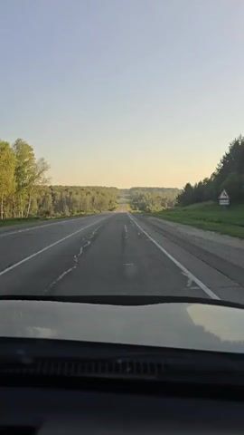 The video shows a view from inside a car as it drives down a paved road lined with trees. The road has white lane markings and appears to be in fair condition with some cracks visible. The sky is clear and blue, indicating daylight. The landscape is green and hilly, suggesting a rural or suburban setting. The camera perspective is from the driver's seat, looking forward through the windshield. There are no people visible inside or outside the car. The sun appears to be low in the sky, casting a warm light. The car is moving at a moderate speed, as indicated by the steady progress down the road.
