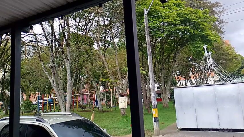 The video opens with a view through an opening, possibly a window or porch, looking towards a green park area with numerous trees and playground equipment. A silver car is partially visible in the foreground, next to a large white metal box and a utility pole. A white, star-topped structure, possibly a festive decoration, is also visible. The sky is overcast.

The camera then pans right, revealing a quiet street lined with two-story buildings of varying colors, including green, white, orange, and light brown. Power lines are visible overhead. The prominent green building on the corner features the name "DENTALIS" with a directional arrow and other signs on its facade. Barred windows and small balconies are common on the buildings. A woman walks away from the camera on the sidewalk, and a motorcycle is parked on the right side of the street.

The camera continues to pan left, showing the same street and buildings again, with the "DENTALIS" sign visible once more. The street appears to be empty of vehicles. The scene depicts an urban setting in Rionegro, Colombia, with a mix of residential or commercial buildings adjacent to a park, under an overcast sky.