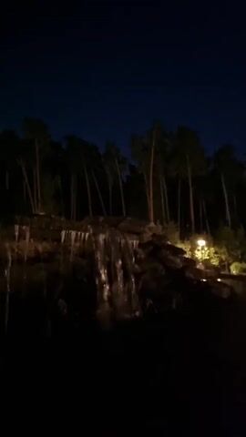A nighttime video shows a waterfall cascading down a rocky incline, surrounded by tall, dark trees. A single light fixture illuminates the rocks to the right of the waterfall, casting a warm glow on the scene. The sky above is a deep, dark blue, indicating that it is night. The video is shaky and the lighting is dim, making it difficult to discern fine details.