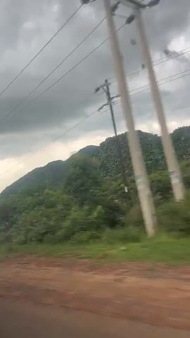 The video shows a view from a moving vehicle traveling along a dirt road next to a green, hilly landscape. The sky is overcast with gray clouds. There are power lines visible across the top of the frame.