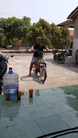 A man is sitting on a white dirt bike. He is wearing a black t-shirt, blue shorts, and a white baseball cap. The man is looking at the camera and smiling. He is in front of a building with trees in the background. There are two other motorcycles parked behind him. There is a large water jug and three glasses on a table in the foreground. The lighting suggests it is daytime.