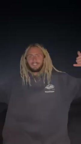 Here is a concise description of the video:

A person with long, blonde dreadlocks stands outside in the dark. They are wearing a gray hoodie with a small logo and are smiling. The person makes hand gestures, holding up a "shaka" sign with each hand. The background is very dark, possibly night, with some vague shapes visible in the distance. Sounds of crashing waves can be heard.