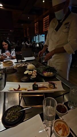 The video shows a chef in a restaurant preparing food on a teppanyaki grill. The chef, wearing a white uniform and a face mask, is chopping vegetables with a spatula and a knife. In front of him, there is a variety of ingredients including sushi rolls, mushrooms, zucchini slices, shredded cabbage, and other vegetables, arranged on plates. There are also small bowls with sauces and condiments around the grill. A woman is seated at the table, looking at her phone. Other diners are visible in the background. The restaurant has a dimly lit interior with decorative wood panels.