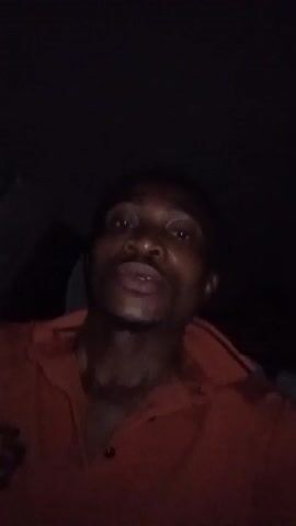 The video captures a dimly lit scene inside a vehicle at night in Iyara, Nigeria. Multiple individuals are present, though mostly obscured by darkness. One person is clearly visible holding an iPhone and possibly a cigarette. Another person's hands are seen holding a red cup. The video begins with a blurry shot of a person's face, then focuses on hands holding objects. Conversations are audible, including repeated words like "TV" and "Malumati." The overall setting suggests people are traveling or gathered in a car during nighttime hours.