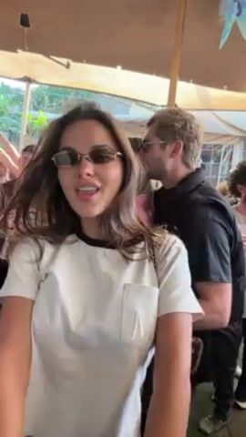 A woman with long brown hair and sunglasses is dancing in a crowd of people under a tent. She wears a white t-shirt with black trim. Behind her are other people dancing, some with drinks. The tent is open-sided, and sunlight streams through.