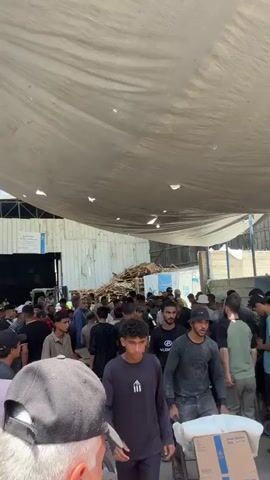 The video shows a crowded indoor space with a large group of people. The area appears to be a warehouse or a large open-air structure with a corrugated metal roof supported by beams. The roof has several holes in it, allowing sunlight to stream through in some areas. The walls are also made of corrugated metal. In the background, there are stacks of wooden pallets. The floor is a mix of dirt and possibly concrete.

The crowd consists primarily of men and boys of various ages. Many are dressed in casual clothing, including t-shirts, long-sleeved shirts, and hats. Some individuals appear to be carrying or pushing carts, suggesting a distribution or collection point for goods. There is a sign on the wall with Arabic writing and a blue and white logo that resembles the United Nations emblem.

The people are standing close together, some in conversation, others looking around. The overall impression is one of activity and possibly a queuing or gathering situation. The lighting is natural, coming from the holes in the roof and possibly an open entrance in the background.
