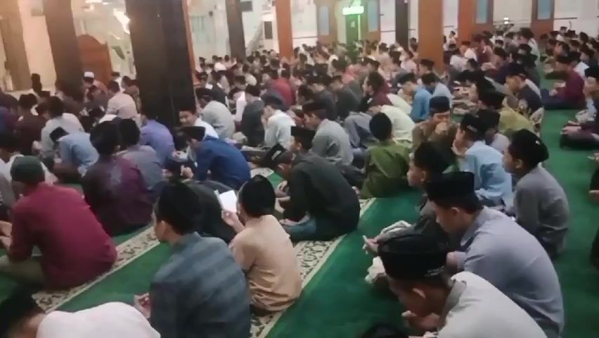 The video shows a large group of people, mostly young males wearing caps, sitting on a green carpeted floor, likely inside a mosque. They are facing in the same direction, suggesting they are participating in a religious gathering or activity. Some individuals are holding books, and their postures indicate attentiveness and reverence. The environment appears to be indoors, with muted lighting, and hints of architectural elements like pillars and windows can be observed in the background. The overall atmosphere seems to be one of communal worship or learning.