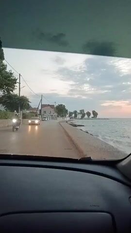 The video shows a view from inside a car driving along a coastal road at dusk. The road runs parallel to the sea on the right side of the frame. On the left side of the road, there are houses and trees. The sky is cloudy and the sun appears to be setting, casting a warm glow on the horizon. There are a few other cars on the road, and the headlights of an approaching car are visible. The water in the sea is choppy. The interior of the car is visible in the foreground, including the dashboard and part of the windshield.