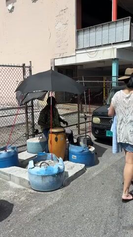 A man is playing a conga drum and smaller drums on the side of a street in what appears to be a warm climate. He is under an umbrella for shade, set up between a building and a chain-link fence. The fence encloses a parking area with parked vehicles. He is sitting on what appears to be a blue bucket or barrel. There are other blue barrels with cement, gravel, or shells in them. The man is wearing a patterned shirt, dark jeans, and dark footwear. He is playing with his hands. A woman walks by in the foreground.