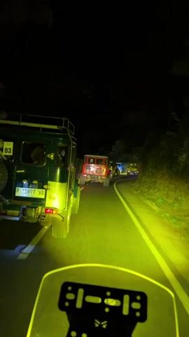 This video captures a convoy of off-road vehicles, specifically what appear to be modified Jeeps or similar rugged SUVs, driving on a winding road at night. The scene is illuminated by the bright, yellow headlights of the vehicles, creating a dramatic and somewhat surreal atmosphere. The road is paved and marked with a white line, suggesting it's a public road, possibly in a mountainous or forested area given the dark, dense foliage visible on the sides of the road.

The perspective is from a vehicle within the convoy, looking forward. In the foreground, the dashboard or handlebars of the leading vehicle are visible, along with its bright yellow headlights casting a wide beam. Directly ahead, the rear of a dark green off-road vehicle is prominent, its license plate clearly visible as "P 1897 VB". Behind this green vehicle, several other vehicles in similar off-road style are following, including a distinctive red one. The procession moves along the curved road, disappearing into the darkness ahead.

The time of day is clearly night, with no natural light sources apparent beyond the vehicle headlights. The weather appears clear, as the darkness is deep and there's no indication of rain or fog. The location context provided is Tosari, Indonesia, which is known for its mountainous terrain and off-road tourism, making this scene plausible. The overall mood conveyed is one of adventure and exploration, a nighttime journey through a scenic, albeit dark, landscape. The glowing headlights cutting through the darkness suggest a determined progression, perhaps a safari or a trek to a specific destination. There is no visible text other than the license plate on the green vehicle.