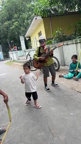 A man wearing a white hat, a green shirt, and gray shorts plays a guitar in the middle of a paved street. A young girl in a striped shirt and black shorts dances in front of him. To the right, another girl in a green tracksuit sits on the ground holding a green object. A bicycle is parked behind the man. A boy in a blue, red, and white jersey stands to the left, pulling a rope that is tied around his waist, facing the young girl who is dancing. The scene appears to be in a residential area with a yellow house and trees in the background.