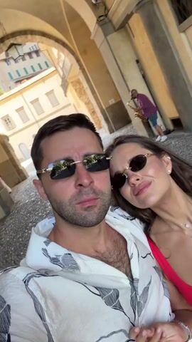 This is a selfie-style video of a couple posing outdoors. The man in the foreground, wearing a white patterned shirt and sunglasses, is looking at the camera. Beside him, a woman in a red top and sunglasses is also looking at the camera, sticking her tongue out and making a rock-and-roll hand gesture. In the background, under an archway, a man in a purple shirt is playing a saxophone. Other people are visible walking in the distance. The scene appears to be a sunny day, with warm lighting indicating it's likely daytime. The architecture suggests a European setting, possibly Italy, with buildings featuring stone and arched doorways. The overall mood is playful and energetic, with the couple appearing happy and engaged. There is no visible text in the video.