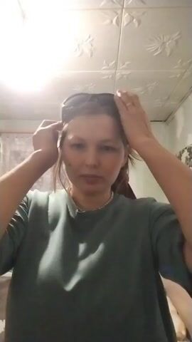 A person, presumably a woman, is putting on sunglasses. She is wearing a green shirt and a necklace. The room appears to be a bathroom or small room, judging by the ceiling tile pattern and glimpse of a curtained window. She smiles briefly while adjusting the sunglasses.