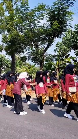 A group of young people, likely students, are walking in a procession down a paved road. They are dressed in matching outfits consisting of red tops, black pants or skirts, and patterned fabric wraps around their waists. Many of the individuals are wearing headscarves, some black and some light pink. The group appears to be organized, walking in rows. The environment is outdoors with abundant green trees and vegetation lining the road. The sky is visible and appears to be blue with some clouds. The overall impression is of a school event or parade.
