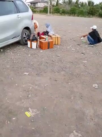 A silver minivan is parked on a gravel surface. Several packages, some wrapped in brown paper and others in white and black, are stacked beside the vehicle. A person wearing a blue hijab is seated among the packages. To the right of the packages, a person in a black shirt and blue jeans, wearing a white cap, is crouched down, seemingly interacting with something on the ground. The environment appears to be outdoors with some vegetation and buildings visible in the background. The ground is covered with loose gravel and some scattered debris.
