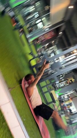 The video shows a man in a gym doing ab exercises while holding his phone. He is lying on a pink yoga mat placed on artificial green turf. The man is wearing a white tank top and black shorts. He has tattoos on his arm. The gym has various exercise machines in the background. The video appears to be shot through a glass window or mirror.
