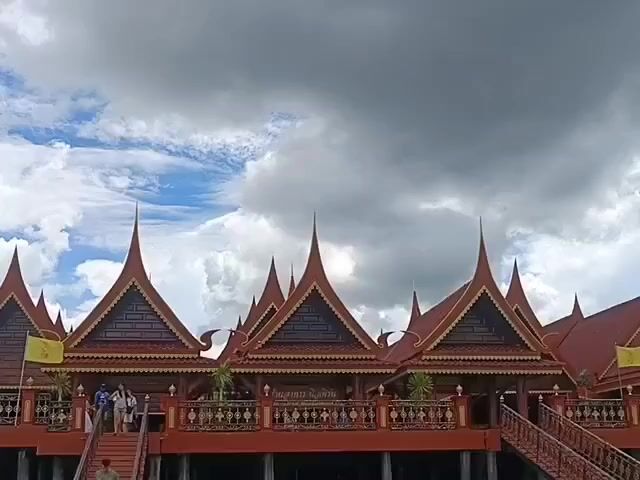 A video shows a traditional Thai building with multiple pointed roofs. There are people on a set of stairs leading up to the building. The sky is cloudy with patches of blue.