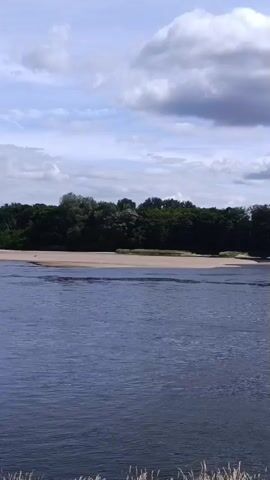 The video shows a wide, calm river under a partly cloudy sky. On the left, a long, narrow boat with a mast and flags is moving across the water. Several people are visible on this boat. To the right of the long boat, a smaller, white motorboat with people on board is also traveling on the river. In the background, a tree-lined shore is visible, with a large white house situated among the trees. The foreground shows a paved walkway and some dry grass.