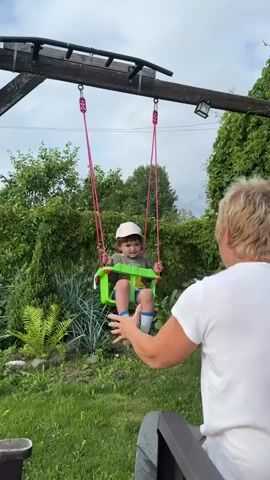 A person with blonde hair is sitting facing a child on a swing. The child is wearing a light-colored hat, a green shirt, and blue shorts. The swing is green and attached to a wooden frame by pink ropes. The person is pushing the swing with their hands, and the child is laughing. The background is a garden with lush green foliage and trees. The sky is cloudy.