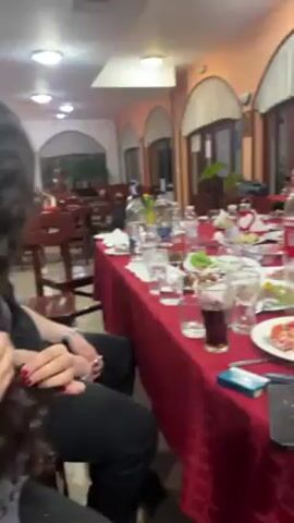 The video shows a group of young men gathered around a long table at what appears to be a restaurant or event hall. The table is covered with a red tablecloth and littered with plates, glasses, bottles, and leftover food, suggesting a recently concluded meal. Several of the men are visible, dressed casually in jeans, t-shirts, and a yellow polo shirt. They are gesturing, showing off, and generally acting jovial, indicating a celebratory or social atmosphere. One man can be seen walking with a hookah hose and holding it as if he's playing the bagpipes. The room is decorated with arched windows, potted plants, and rustic furniture. The lighting is bright, and the overall impression is of a lively, informal gathering.