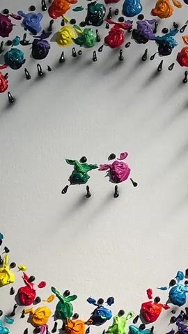 The video displays a piece of art with a white background and a black frame. The art features numerous small, colorful figures made of paint and small black pins. The figures are arranged in a circular pattern around two central figures, one green and one pink, who are also made of paint and pins and appear to be dancing or holding hands. The figures are varied in color, including red, blue, yellow, green, pink, and purple. The lighting casts shadows from the figures and pins onto the white background, creating a textured effect. The camera slowly zooms out, revealing the full artwork and its frame. The artwork is displayed on a light-colored wall.